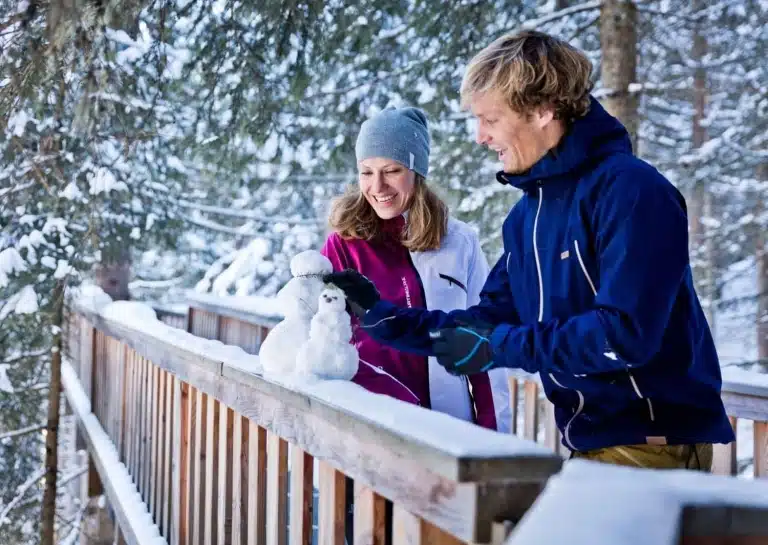 Baumzipfelweg im Winter Paar steht auf Brücke und baut Mini-Schneemann