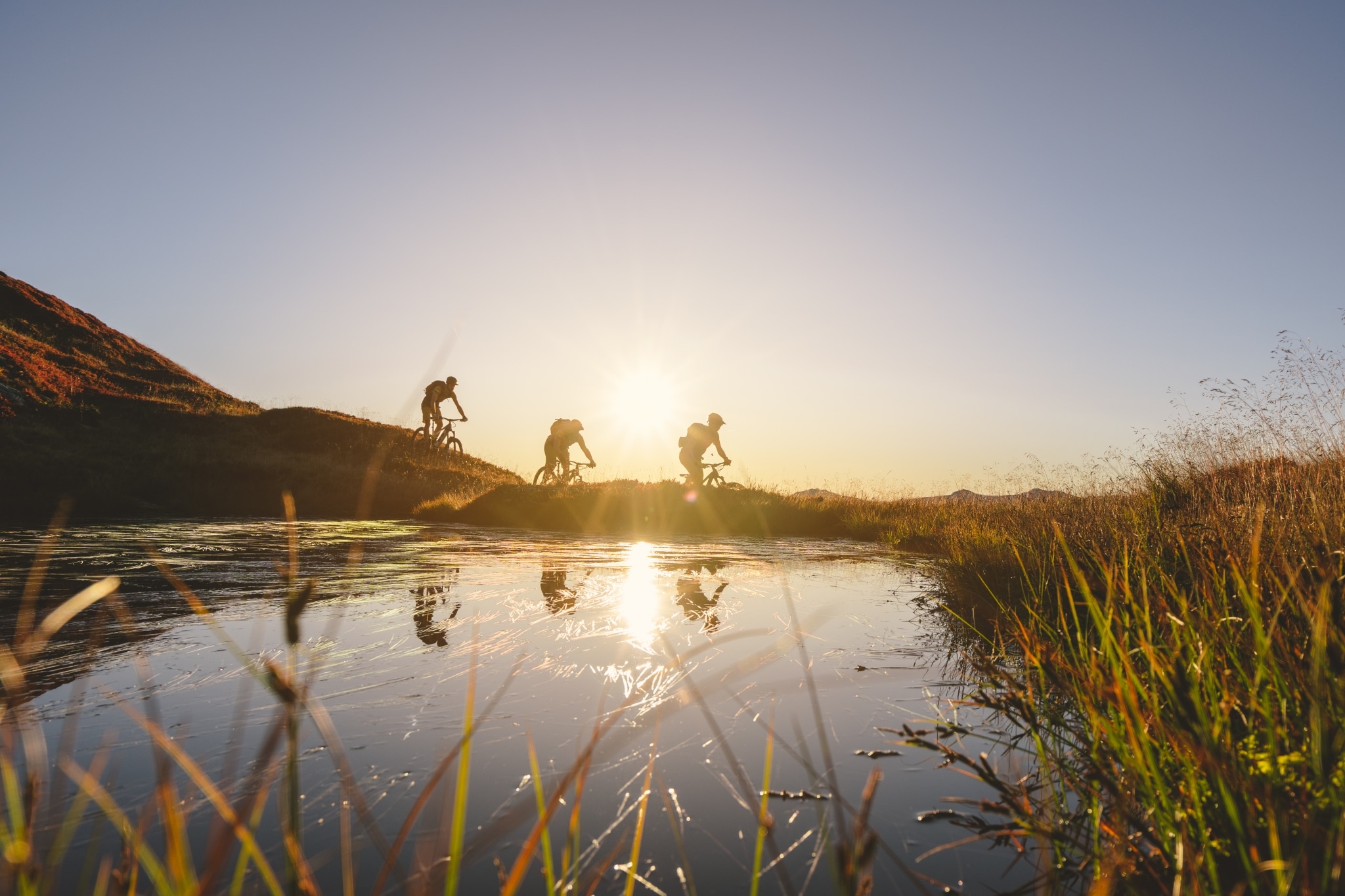 3 Mountainbiker fahren bei Sonnenuntergang Hand hinunter im Vordergrund ist ein See zu erkennen