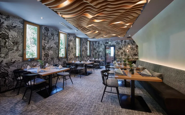 Holzdecke im Restaurant Der Speisesaal mit einem Holzelement auf der Decke sowie weiß-schwarzen Tapeten und einigen gedeckten Tischen.