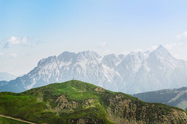 Ein grüner Berg mit Gipfelkreuz im Vordergrund und ein Bergmassiv in der Ferne.