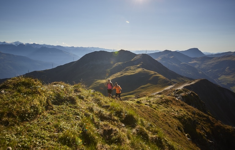 Zwei Wanderer gehen über eine Almwiesen-Kuppe bergaufwärts, im Hintergrund die Salzburger Berwelt