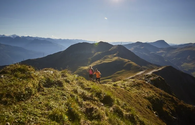Wandern in Saalbach Hinterglemm Zwei Wanderer gehen über eine Almwiesen-Kuppe bergaufwärts, im Hintergrund die Salzburger Berwelt