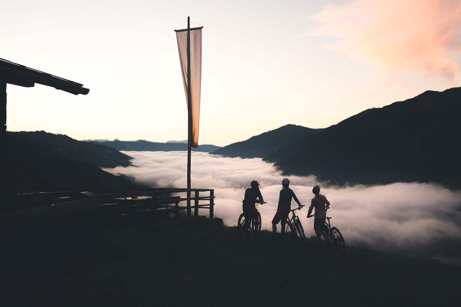 dunkles Bild am Gipfel eines Bergs. Nebelschleier sind im Tal. Drei Biker stehen am Berg mit ihren Bikes